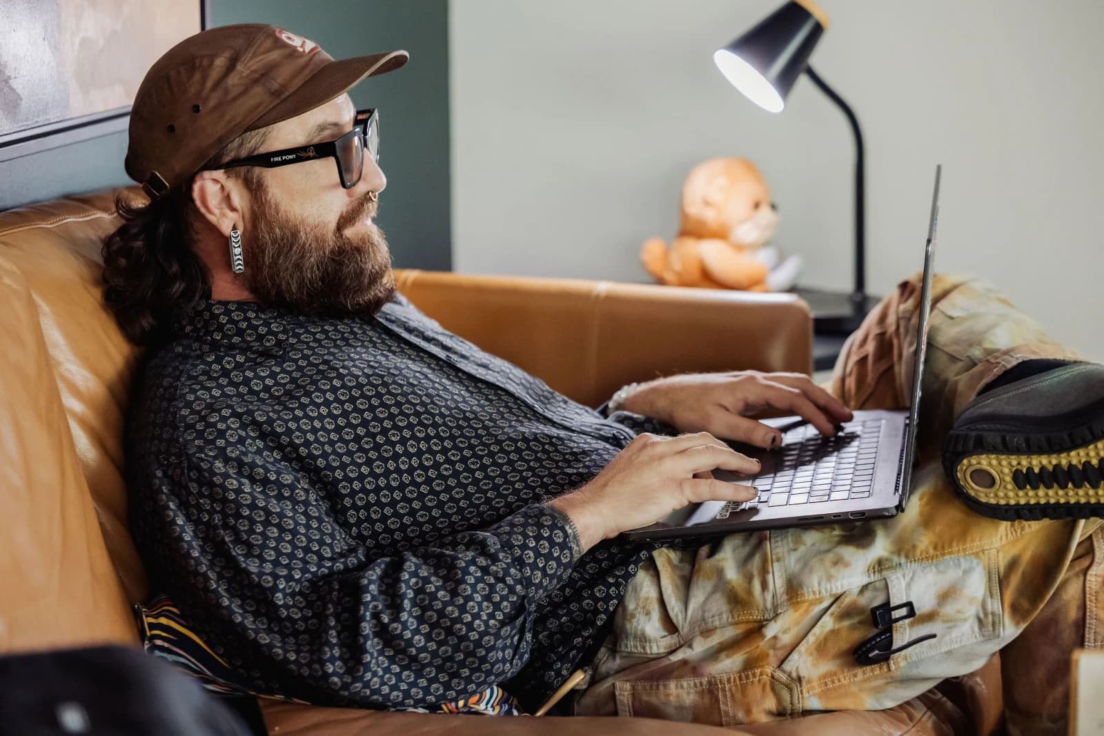 Kris Krüg kicked back on a couch at FUNK, cap and glasses, typing on a laptop with a plush otter and a desk lamp behind him
